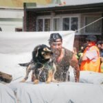 A Shepherds' Shemozzle competitor being sprayed by a wet black and tan dog shaking as they climb over a race obstacle, with an orange-vested official and crowd watching behind an 