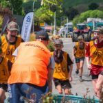 A group of intermediate competitors in numbered vests carrying collars in their mouths, supervised by a person wearing a security vest. Trees, bushes, and a tractor are visible in the background.