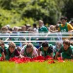 Junior competitors in green bibs crawl under an obstacle with redcaps scattered on the grass, cheered on by a crowd of spectators.