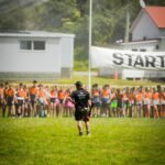 Teen competitors in orange bibs stand lined up on a grass field facing a Shemozzle crew member holding a starter's pistol, with a 